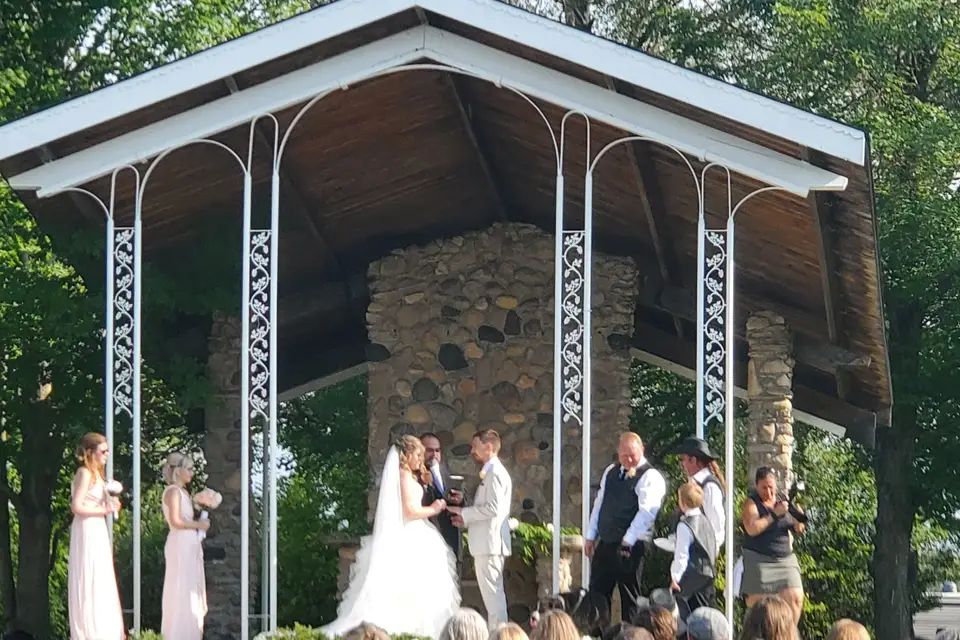An outdoor wedding ceremony under a stone pavilion, with ceremony sound by Hammer DJ Services